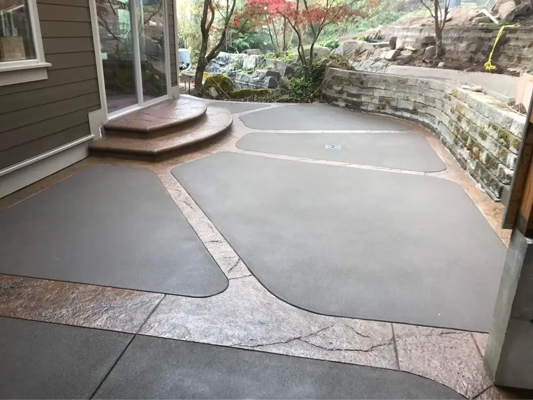 Wide view of a custom two-tone concrete patio in Puyallup with stamped borders and curved steps near a rock wall.